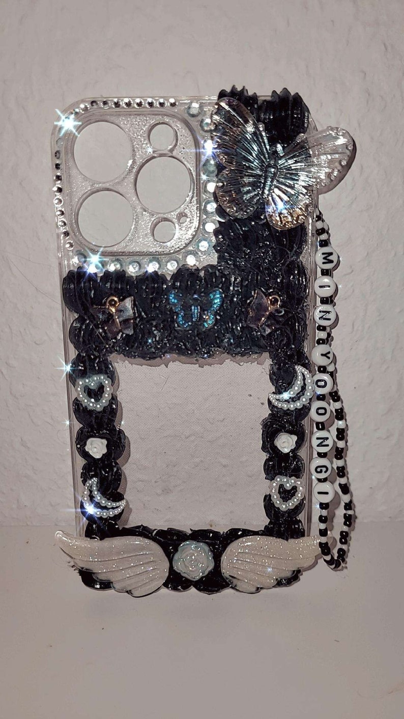 Peut inclure: Une coque de t&eacute;l&eacute;phone transparente avec un motif noir et blanc. La coque est orn&eacute;e d'un charme papillon, d'une cha&icirc;ne de perles et de perles noires, et d'une paire d'ailes blanches. La coque a &eacute;galement un dos transparent avec une finition scintillante.