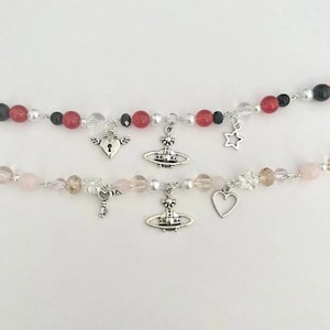 Könnte beinhalten: Zwei Perlenketten mit Anhängern. Die obere Kette hat rote, schwarze und weiße Perlen mit einem silbernen Schloss-, Stern- und Kugelanhänger. Die untere Kette hat rosa, weiße und braune Perlen mit einem silbernen Schlüssel-, Herz- und Kugelanhänger.