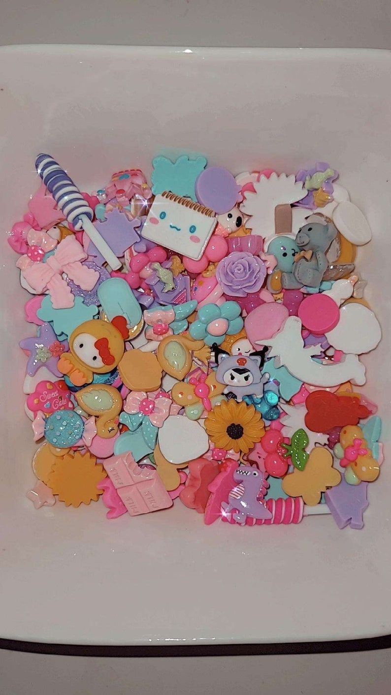 Puede incluir: Una colecci&oacute;n de abalorios de resina coloridos en varias formas, incluyendo lazos, flores y personajes de dibujos animados. Los abalorios son en tonos de rosa, amarillo, azul y morado, con algunos con acentos de purpurina. Tambi&eacute;n se ven una piruleta y un bloc de notas.