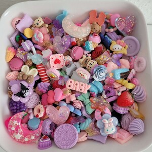 Puede incluir: Una colecci&oacute;n de coloridos dijes de resina en un plato blanco. Los dijes incluyen varias formas como corazones, lazos, osos, flores y dulces. La paleta de colores es predominantemente rosa pastel, morado y azul, con algunos detalles brillantes.
