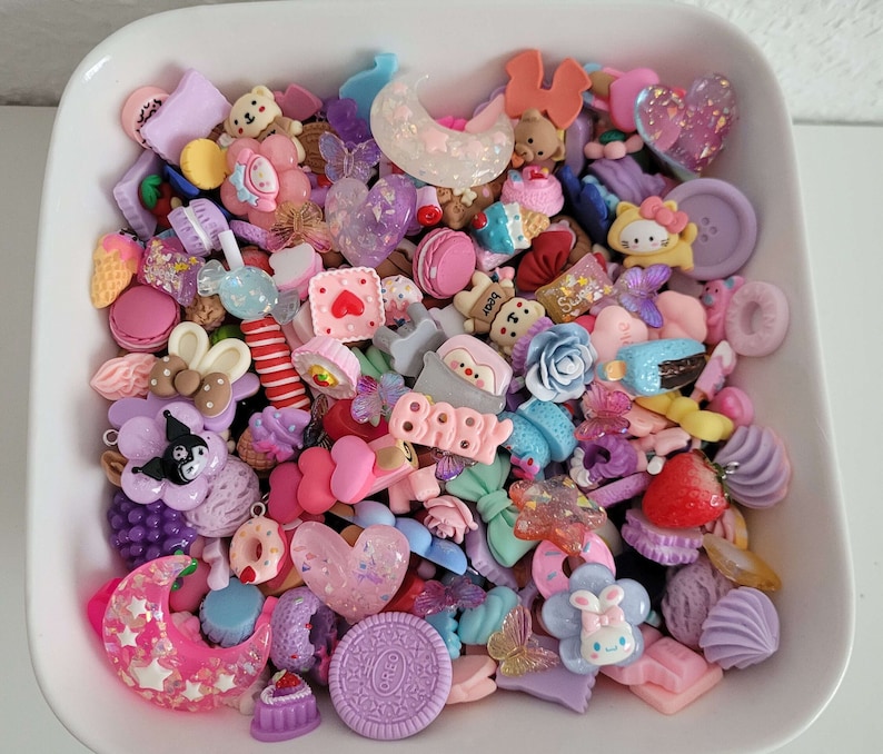 Puede incluir: Una colecci&oacute;n de coloridos dijes de resina en un plato blanco. Los dijes incluyen varias formas como corazones, lazos, osos, flores y dulces. La paleta de colores es predominantemente rosa pastel, morado y azul, con algunos detalles brillantes.
