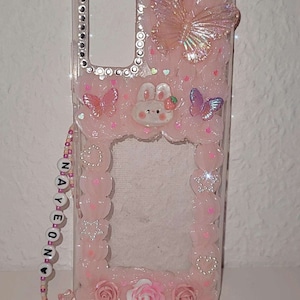 Peut inclure: Une coque de t&eacute;l&eacute;phone rose avec un dos transparent et un motif rose, blanc et argent. La coque est orn&eacute;e d'un papillon, d'un lapin et de fleurs. La coque est &eacute;galement dot&eacute;e d'une sangle perl&eacute;e rose avec le mot "NAYEON" dessus.