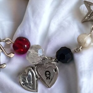 Op de afbeelding: Een zilveren armband met verschillende bedels, waaronder een hartvormig medaillon, een muzieknoten, een ster en kralen in rood, zwart, wit en transparant.