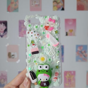 May include: A clear phone case with a green and white background. The case is decorated with 3D charms, including a Hello Kitty character, a frog, a cake, a lollipop, and a cookie. The case also has white and green stars and flowers.