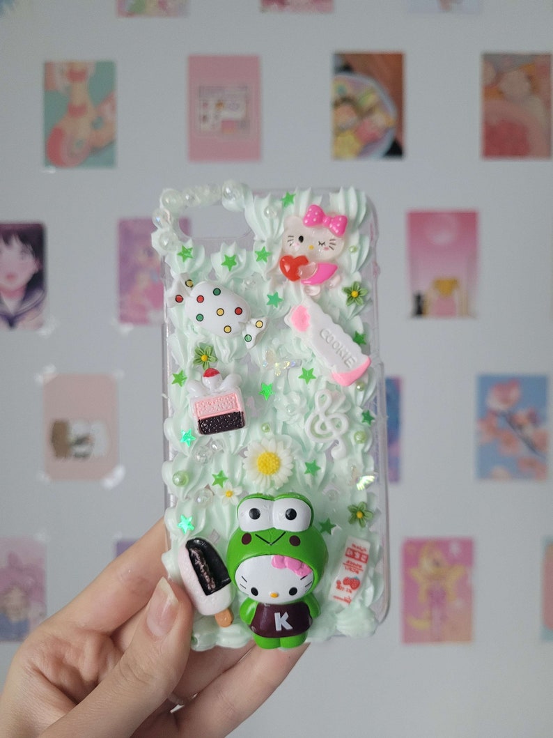 May include: A clear phone case with a green and white background. The case is decorated with 3D charms, including a Hello Kitty character, a frog, a cake, a lollipop, and a cookie. The case also has white and green stars and flowers.