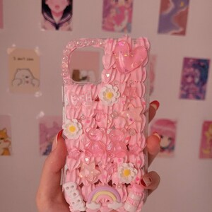 May include: A pink phone case with a 3D design featuring a rainbow, clouds, flowers, stars, and hearts. The case is made of clear plastic with a pink, white, and glitter design.