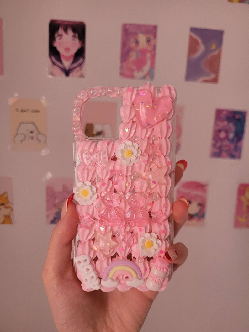 May include: A pink phone case with a 3D design featuring a rainbow, clouds, flowers, stars, and hearts. The case is made of clear plastic with a pink, white, and glitter design.