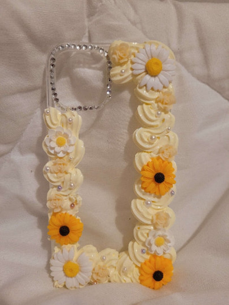 Peut inclure: Une coque de t&eacute;l&eacute;phone transparente d&eacute;cor&eacute;e de fleurs blanches et jaunes. La coque a une bordure scintillante et est orn&eacute;e de perles blanches.