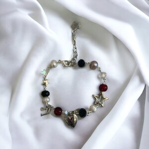 Op de afbeelding: Een zilveren armband met bedels in de vorm van een hart, sterren en een muzieknoten. De armband is gemaakt met zwarte, rode, witte en parelkleurige kralen.