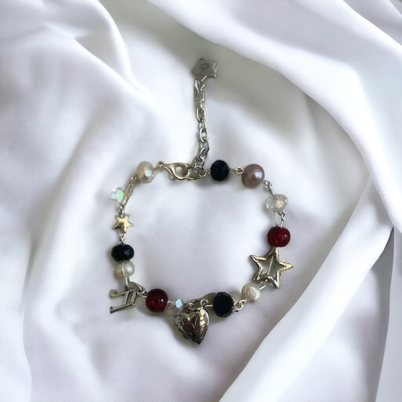 Op de afbeelding: Een zilveren armband met bedels in de vorm van een hart, sterren en een muzieknoten. De armband is gemaakt met zwarte, rode, witte en parelkleurige kralen.