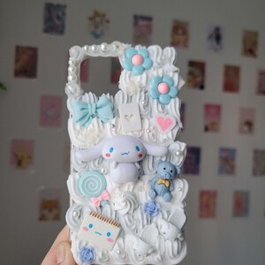 May include: A white phone case decorated with white frosting, blue flowers, a white and blue bow, a white lollipop, a white teddy bear, a white notepad, and a white cartoon character with blue eyes.