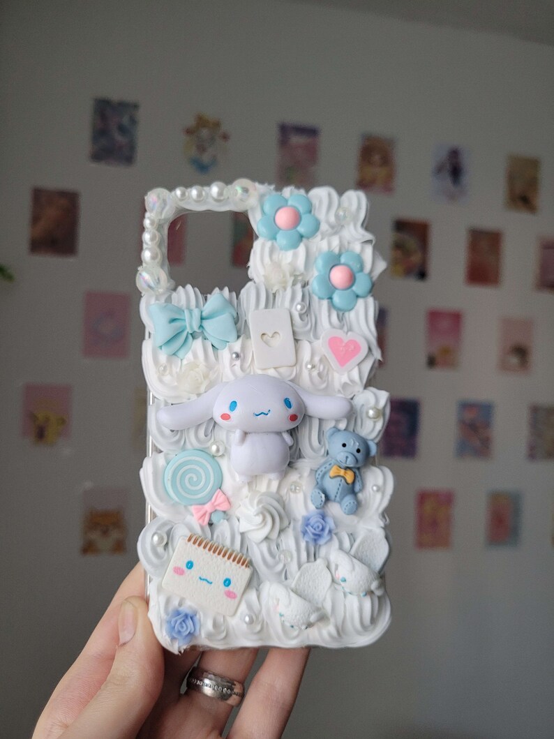 May include: A white phone case decorated with white frosting, blue flowers, a white and blue bow, a white lollipop, a white teddy bear, a white notepad, and a white cartoon character with blue eyes.