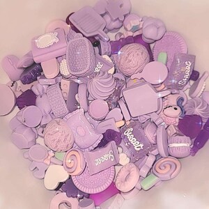 Puede incluir: Una colecci&oacute;n de abalorios de resina de color lavanda con formas variadas, como helados, galletas y caramelos. Algunos abalorios tienen la palabra "Sweet". Los abalorios est&aacute;n en un cuenco blanco.