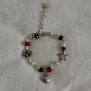 Op de afbeelding: Een zilveren armband met rode, witte en zwarte kralen. De bedels zijn een ster, een hart en een lotusbloem.