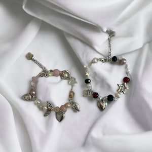 Op de afbeelding: Twee zilveren armbandjes met roze, witte en zwarte kralen. E&eacute;n armbandje heeft een hartbedel, aardbeibedels en engelenvleugelbedels. Het andere armbandje heeft een hartbedel, sterbedels en een muzieknootbedel.