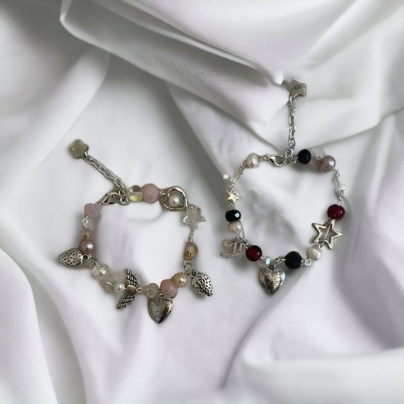 Op de afbeelding: Twee zilveren armbandjes met roze, witte en zwarte kralen. E&eacute;n armbandje heeft een hartbedel, aardbeibedels en engelenvleugelbedels. Het andere armbandje heeft een hartbedel, sterbedels en een muzieknootbedel.