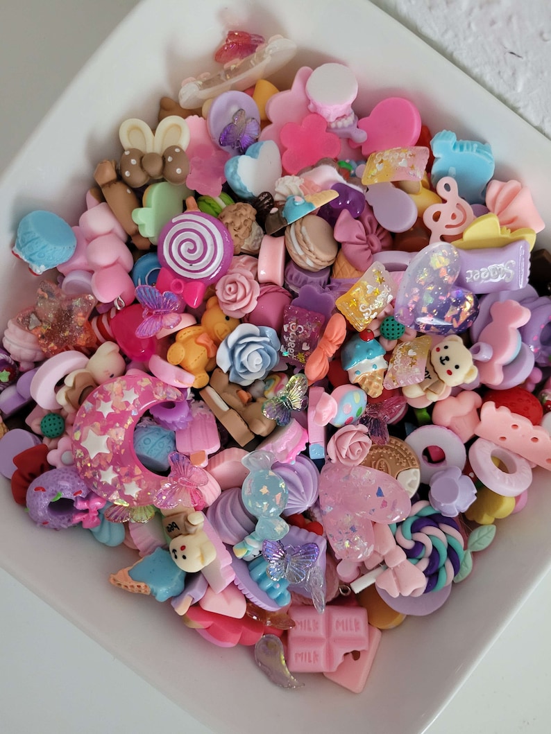 Puede incluir: Un cuenco blanco lleno de una variedad de abalorios de resina coloridos. Los abalorios incluyen piruletas, osos, lazos, corazones y otros dulces en tonos de rosa, morado y azul. Algunos abalorios tienen detalles de purpurina.