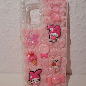 May include: A pink phone case with a 3D design featuring a pink and white Hello Kitty character, a pink lollipop, a pink ice cream cone, a pink bear, and a pink bow. The case is decorated with white pearls around the edges.