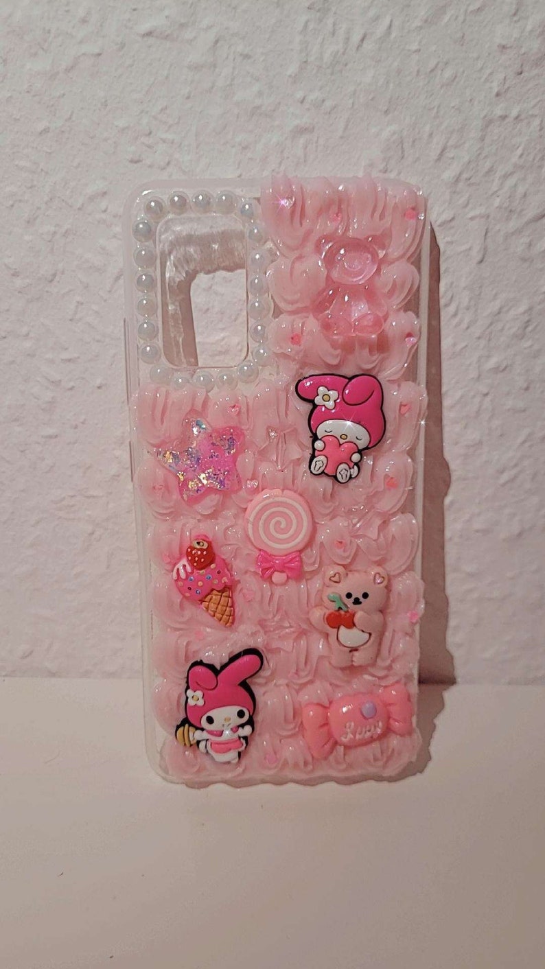 May include: A pink phone case with a 3D design featuring a pink and white Hello Kitty character, a pink lollipop, a pink ice cream cone, a pink bear, and a pink bow. The case is decorated with white pearls around the edges.