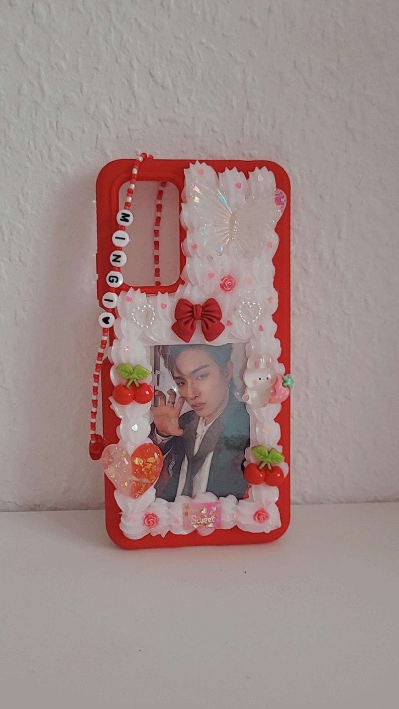Peut inclure: Coque de t&eacute;l&eacute;phone rouge avec un motif floral blanc et rose, une photo d'une personne et un charme perl&eacute; avec le mot "MINGI" dessus.