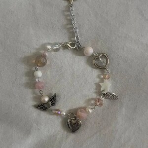 Op de afbeelding: Een delicate armband met een zilveren ketting en roze en witte kralen. De armband heeft bedels in de vorm van harten, sterren en vleugels.