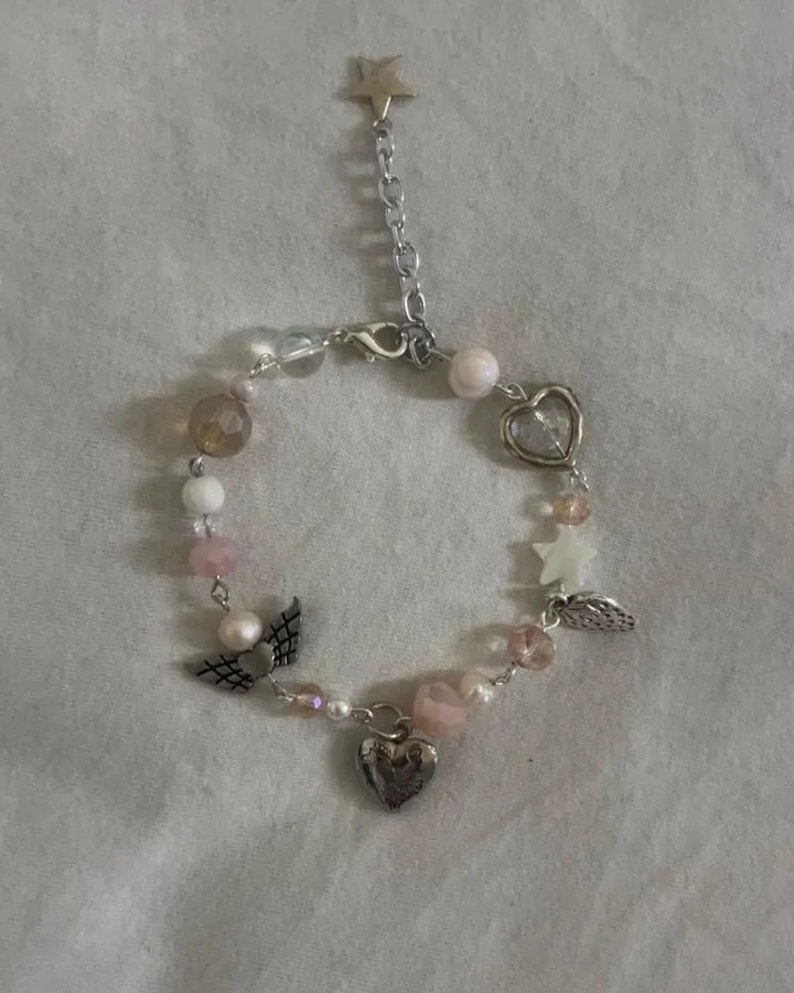 Op de afbeelding: Een delicate armband met een zilveren ketting en roze en witte kralen. De armband heeft bedels in de vorm van harten, sterren en vleugels.