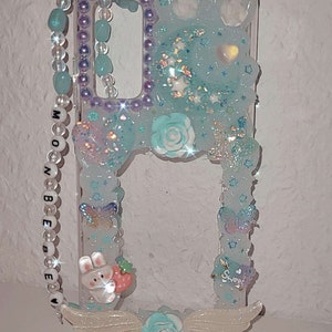 Peut inclure: Une coque de t&eacute;l&eacute;phone transparente avec un fond bleu clair et des paillettes. La coque pr&eacute;sente un lapin blanc avec une fraise rouge, une fleur blanche et des ailes blanches. La coque a &eacute;galement une sangle perl&eacute;e avec les mots "Moonbeam" dessus.