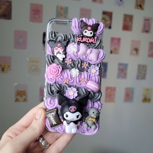 May include: A purple and black phone case with a 3D design featuring a black and white cartoon character, flowers, and a chocolate bar with the word "Sweet" on it. The character is wearing a bow and has a skull on its head. The case also has the word "Kuromi" on it.