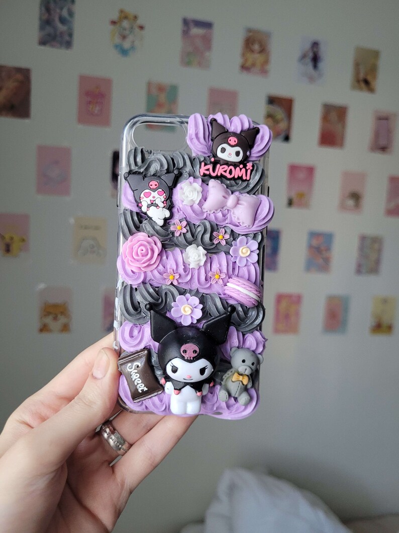 May include: A purple and black phone case with a 3D design featuring a black and white cartoon character, flowers, and a chocolate bar with the word "Sweet" on it. The character is wearing a bow and has a skull on its head. The case also has the word "Kuromi" on it.