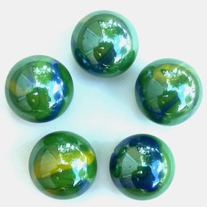 Pode incluir: Cinco bolas de gude de vidro verde com redemoinhos azuis e amarelos dispostas sobre uma superfície branca. As bolas de gude têm um acabamento brilhante, refletindo a luz e o ambiente. Estas bolas de gude são um brinquedo clássico.