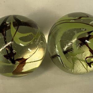 May include: Two green glass marbles with brown and white swirls. The marbles are opaque and have a glossy finish.