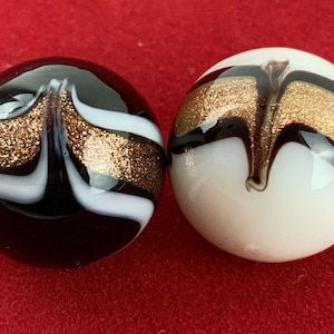 May include: Two decorative glass marbles. One is black with white and gold glitter swirls. The other is white with black and gold glitter swirls. The marbles have a glossy finish and are set against a red background.