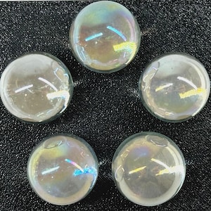 May include: Five iridescent glass marbles with a clear, reflective surface. The marbles have a circular shape and display a range of colors, including blue, yellow, and green. They are arranged on a black, textured background.