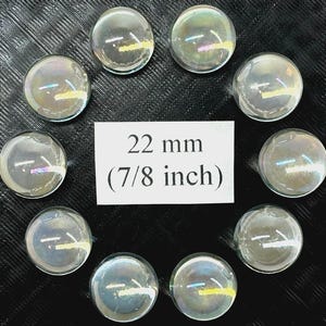 Può includere: Una collezione di dieci biglie di vetro trasparente e iridescente, ciascuna di circa 22 mm di diametro, disposte a cerchio. Un cartoncino bianco mostra la dimensione delle biglie, pari a 7/8 di pollice. Le biglie hanno una lucentezza riflettente simile all'arcobaleno.