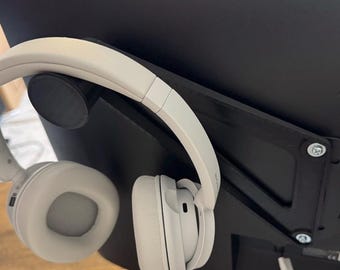 Op de afbeelding: Een paar lichtgrijze over-ear hoofdtelefoons hangen aan een zwarte hoofdtelefoonhouder die aan een zwarte computermonitor is bevestigd. De draadloze hoofdtelefoon heeft een gewatteerde hoofdband en oorschelpen.
