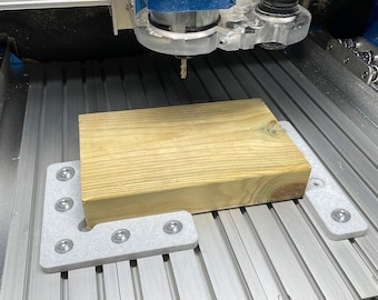 May include: A close-up of a wooden block being cut by a CNC router. The block is secured to a gray L-shaped bracket with screws. The router is positioned above the block, ready to cut.