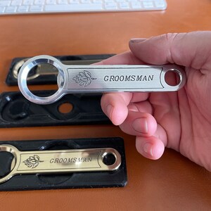 Puede incluir: Dos abridores de botellas plateados con un diseño negro y dorado. Los abridores tienen un diseño de hoja de lúpulo y la palabra "GROOMSMAN" grabada en ellos.