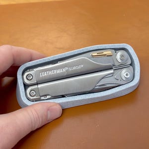以下が含まれることがあります： ライトブルーのケースに入ったLeatherman Surgeマルチツール。マルチツールはシルバーで、「LEATHERMAN SURGE」の文字が側面に印刷されています。ケースは角が丸い長方形です。