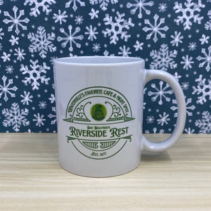 May include: White ceramic mug with a curved handle. The mug features green text that reads "Waterville's Favorite Cafe & Night Spot" and "Riverside Rest" with a frog graphic. The text "Est. 1977" is also printed on the mug.