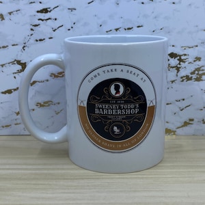 May include: White ceramic mug with a black and gold design featuring the text "Come take a seat at Sweeney Todd's Barbershop, Fleet Street, London. Est. 1846. No closer shave in all of London."