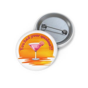 May include: A round pin-back button featuring a graphic of a pink cocktail in a martini glass with a citrus slice garnish, set against an orange sunset background with the text You had your qualms?? and a metal pin mechanism on the back.