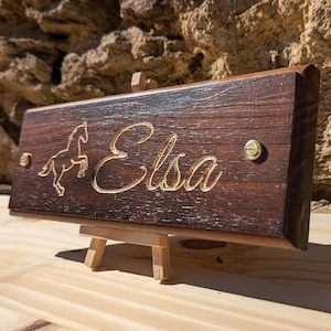 Può includere: Un cartello in legno marrone scuro con il nome "Elsa" inciso in lettere marroni chiare. Una piccola sagoma di cavallo è anche incisa sul cartello. Il cartello è su un piccolo cavalletto in legno.