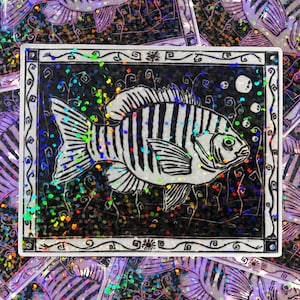 May include: A black and white striped fish sticker with a decorative border. The fish is the focal point, with bubbles and a dark background. The sticker has a holographic, glittery effect, and is surrounded by other similar stickers.
