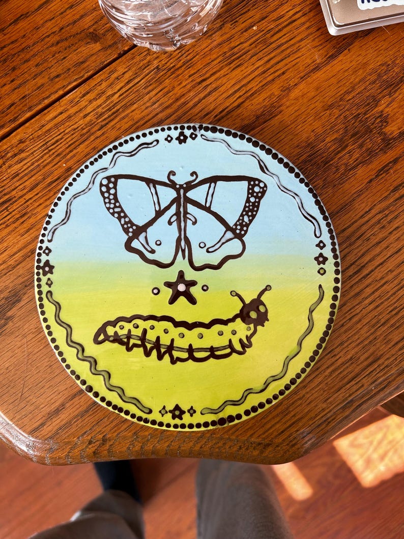 K&ouml;nnte beinhalten: Ein runder Untersetzer aus Keramik mit schwarzem Rand und einem blauen und gr&uuml;nen Farbverlauf. Auf dem blauen Teil ist ein schwarzer Schmetterling gemalt, auf dem gr&uuml;nen Teil eine schwarze Raupe. Zwischen Schmetterling und Raupe ist ein wei&szlig;er Stern gemalt.