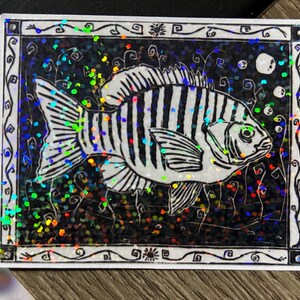 May include: A rectangular sticker featuring a black and white illustration of a fish with striped markings. The fish is set against a dark background with iridescent glitter. The sticker has a decorative white border.