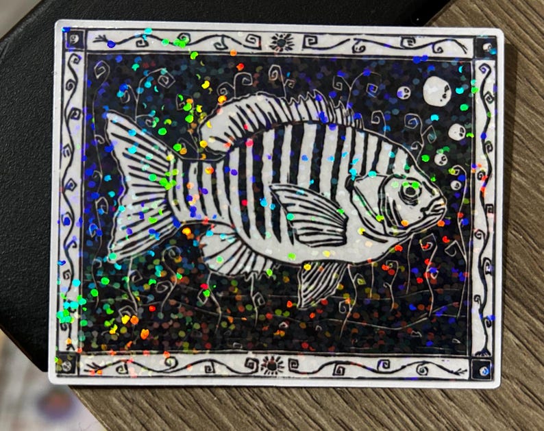 May include: A rectangular sticker featuring a black and white illustration of a fish with striped markings. The fish is set against a dark background with iridescent glitter. The sticker has a decorative white border.
