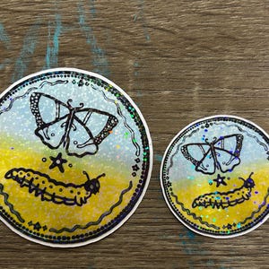 K&ouml;nnte beinhalten: Zwei runde Aufkleber mit holografischem Finish. Der gr&ouml;&szlig;ere Aufkleber zeigt einen schwarz-wei&szlig;en Schmetterling mit einem Stern und einer Raupe. Der kleinere Aufkleber zeigt einen schwarz-wei&szlig;en Schmetterling mit einem Stern und einer Raupe.