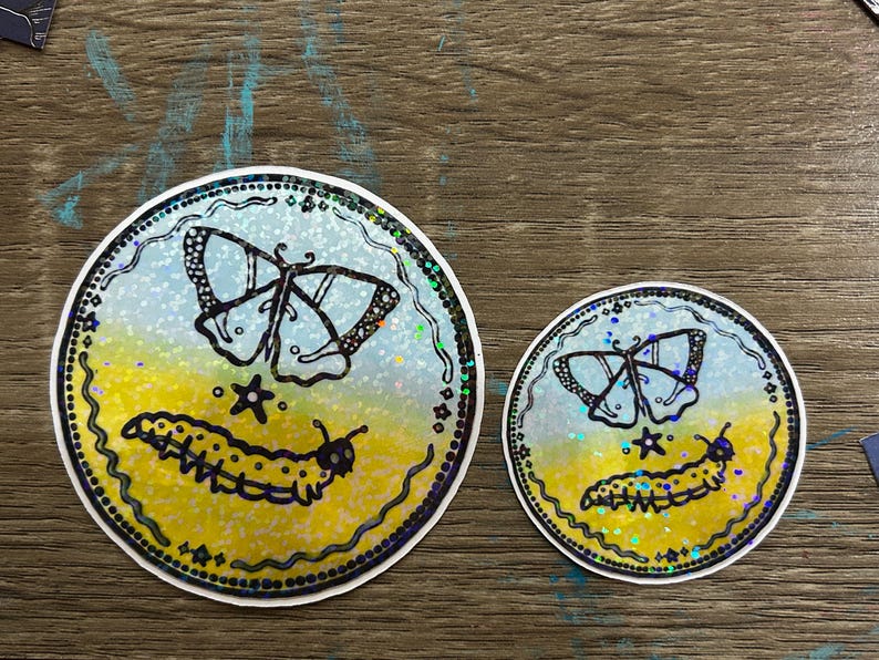 K&ouml;nnte beinhalten: Zwei runde Aufkleber mit holografischem Finish. Der gr&ouml;&szlig;ere Aufkleber zeigt einen schwarz-wei&szlig;en Schmetterling mit einem Stern und einer Raupe. Der kleinere Aufkleber zeigt einen schwarz-wei&szlig;en Schmetterling mit einem Stern und einer Raupe.