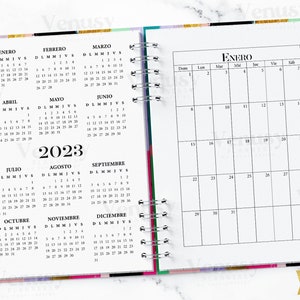 Puede incluir: Un planificador con espiral abierto a un calendario de 2023. La p&aacute;gina izquierda muestra un calendario anual en espa&ntilde;ol, mientras que la p&aacute;gina derecha muestra un calendario mensual de enero. Se ve un bol&iacute;grafo y un clip dorados.
