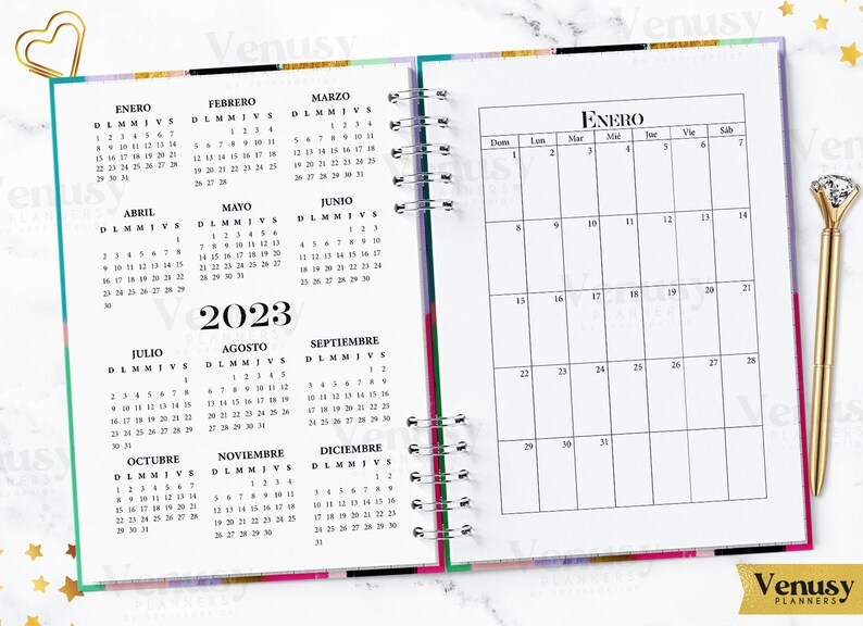 Puede incluir: Un planificador con espiral abierto a un calendario de 2023. La p&aacute;gina izquierda muestra un calendario anual en espa&ntilde;ol, mientras que la p&aacute;gina derecha muestra un calendario mensual de enero. Se ve un bol&iacute;grafo y un clip dorados.